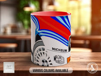 A colorful ceramic mug with a design featuring a car and the text "MICHELIN" and "MAGNETI MARELLI" printed on it. The mug is placed on a wooden surface, and there is text indicating that various colors are available.