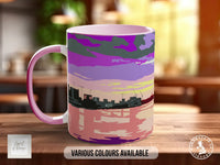 A pink and purple mug with a colorful cityscape design on it, sitting on a wooden surface.