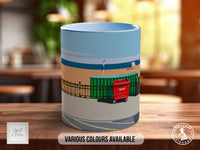 A ceramic mug with a colorful illustration depicting a shipping container, a fence, and a boat on a body of water.