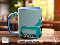 A white and blue ceramic mug with the word "MAXELL" printed on it, sitting on a wooden surface.