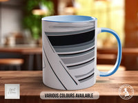 A white mug with a blue handle, featuring a black and gray striped design, sits on a wooden surface.
