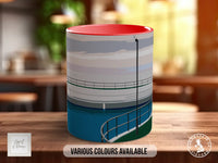 A cylindrical mug with a red rim and a design depicting a body of water, possibly a lake or a river, is displayed on a wooden surface.