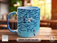 A blue mug with a water-themed design, featuring a person standing in the water and another person standing on the shore. The mug is placed on a wooden surface, and there is text indicating that various colors are available.
