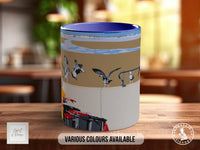 A ceramic mug with a beach scene design, featuring seagulls and a boat. The mug is placed on a wooden surface, and there is text indicating that various colors are available.